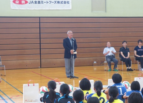 前に立ちあいさつの言葉を述べる三郷市長と、各列の前にバレーボールと紙袋が置かれ、その後ろに整列して体育座りをし話を聞く子どもたちの後方から写した写真