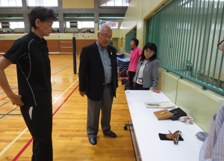 体育館の端に設置された白いテーブルの上に3個のメダルが置いてあり、三郷市長と関係者がメダルを鑑賞している様子の写真
