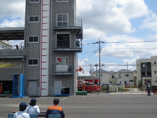 屋外に設置された長机の席に座った消防職員の方が見守るなか、高さが表示されたコンクリート造りの塔で作業着を着用した消防職員の方々が高所からロープで降りている訓練の様子の写真