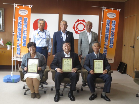 国旗と三郷市旗が飾られた室内の左右にのぼり旗が設置され、前列の椅子に指定書を交付された3名の方が座り、後列に市長と消防職員の男性、スーツ姿の男性が並んでいる写真