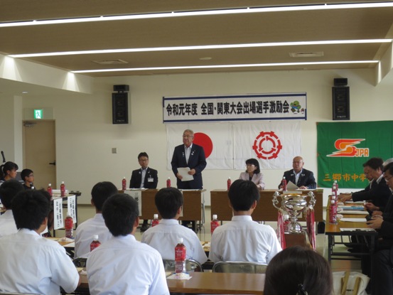 「令和元年度全国・関東大会出場選手激励会」と書かれた横断幕や国旗と三郷市旗が飾られた室内に、関係者や子供たちが座っている長机の席が向かい合わせで設置され、右側の席の前にトロフィーが並び、前の席から立ち上がった市長が挨拶を行っている様子の写真