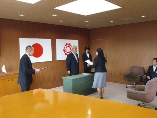 国旗と三郷市旗が飾られた室内で、薄黄緑色のクロスがかけられた小さな台を挟んで立っている市長が椎貝委員に任命書を交付した様子の写真