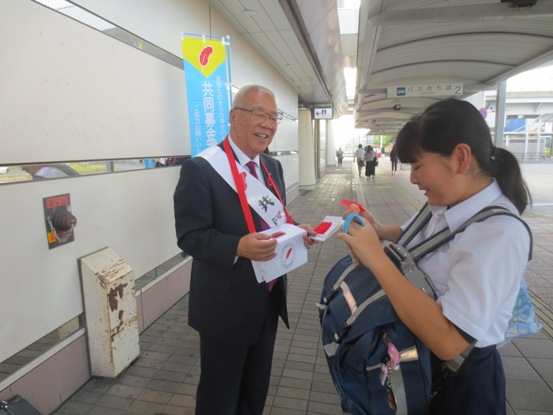 駅の通路に設置されたのぼり旗の手前で募金箱を首にかけた市長が募金に協力してくれた女子生徒に笑顔で赤い羽根を渡している写真