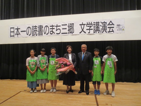 「日本一の読書のまち三郷文学講演会」と書かれた横幕の下、市長と花束を持った林真理子先生を挟んで黄緑色のエプロンをつけた5名の子ども司書の皆さんが横一列に並んでいる写真
