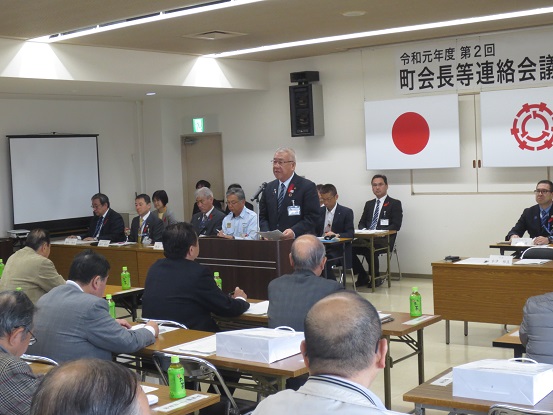 「令和元年度第2回町会長等連絡会議」と書かれた横幕と国旗と三郷市旗が飾られた室内で、関係者と参加者が向かい合って設置された長机の席に座り、中央の演壇で挨拶を行っている市長の写真
