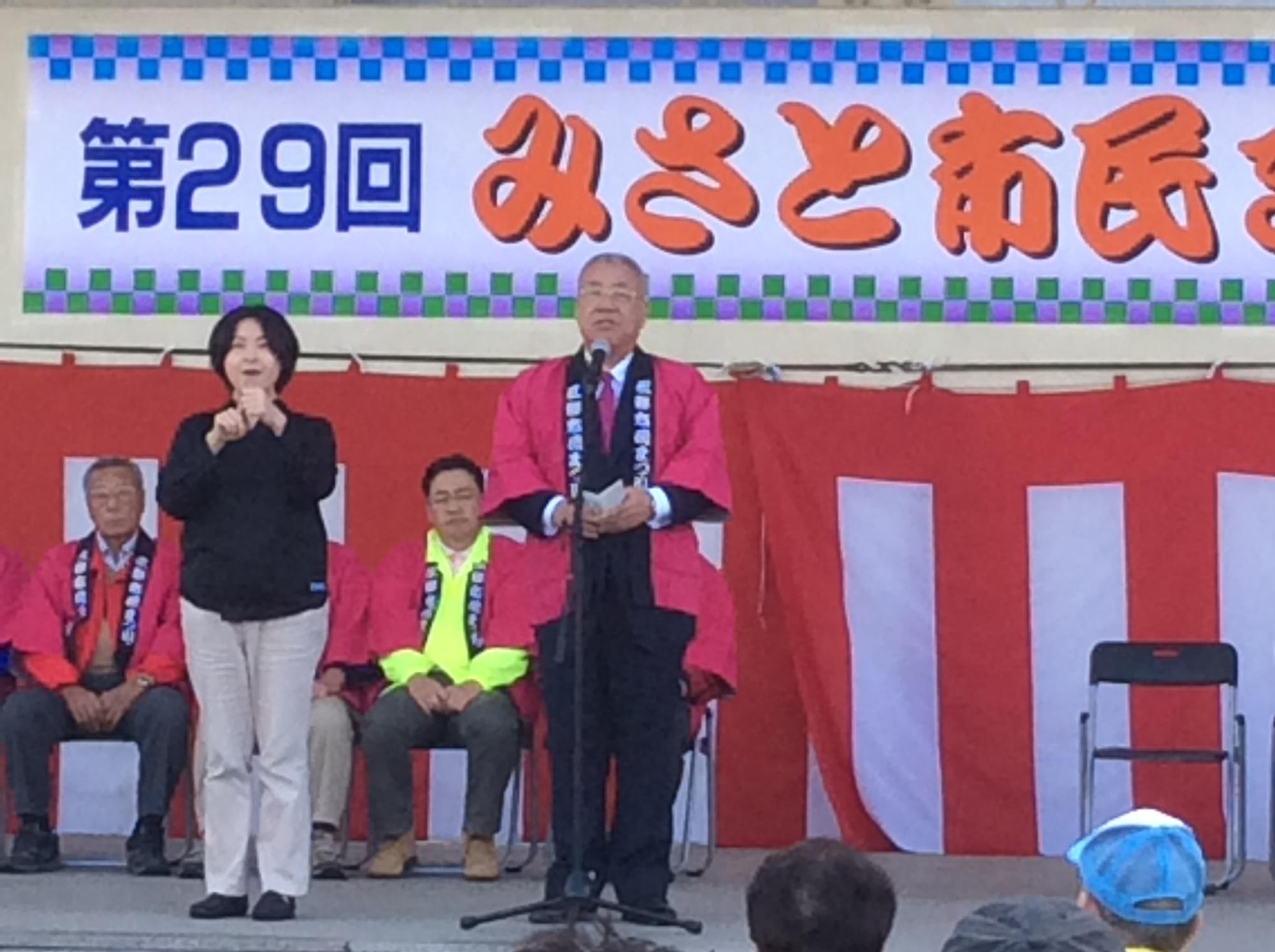 「第29回みさと市民まつり」と書かれた横断幕や紅白幕が設置されたステージの左側に黒色のシャツを着た女性の手話と同時にマイクの前で挨拶を行っている市長の写真