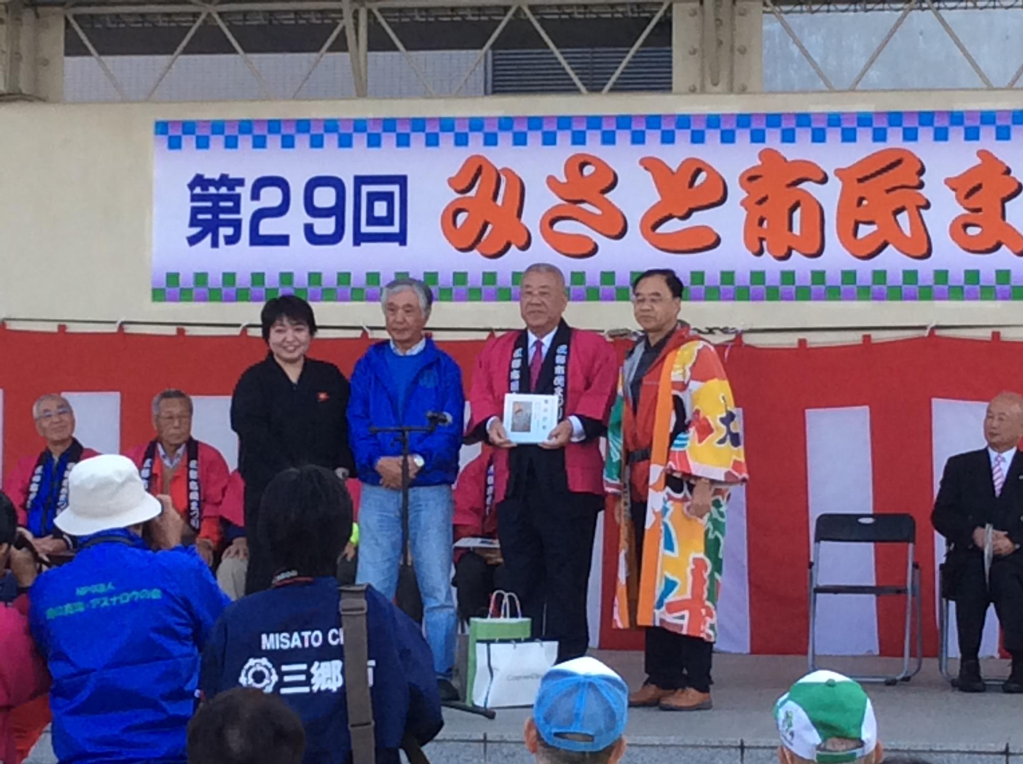 ステージの上で赤色の法被を着た市長が寄贈していただいた本を持ち、左側に男性と女性、右側に男性と一緒に並んで立っている写真