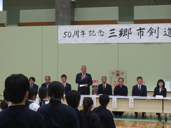 「50周年記念三郷市剣道大会」と書かれた横幕が貼られた体育館に設置された、長机の席に座っている関係者と道着を着用した子供たちが向かい合って立ちながら、中央の席から挨拶を行っている市長の話を聞いている様子の写真
