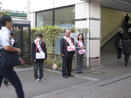 「共同募金」と書かれた白いタスキと赤と白の募金箱をかけた三郷市長と女性、男子学生が新三郷駅の前に立ち、街頭募金活動を行っている様子の写真