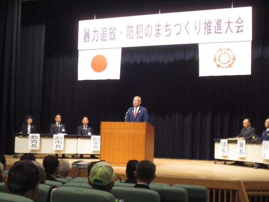 「暴力追放・防犯のまちづくり推進大会」と書かれた横断幕と国旗と三郷市旗が飾られたステージ左右に関係者が座り、中央の演壇に立って挨拶を行っている市長の写真