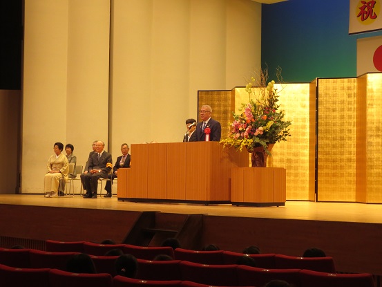 奥に金屏風が立てかけられたステージ左側に関係者が座り、中央の演壇で挨拶を行っている市長の写真