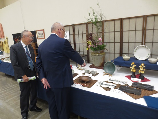 花が生けられた大きな花瓶や皿などの作品が展示され、中央の2つのコーヒーカップを指さして見ている男性と市長の後ろ姿の写真
