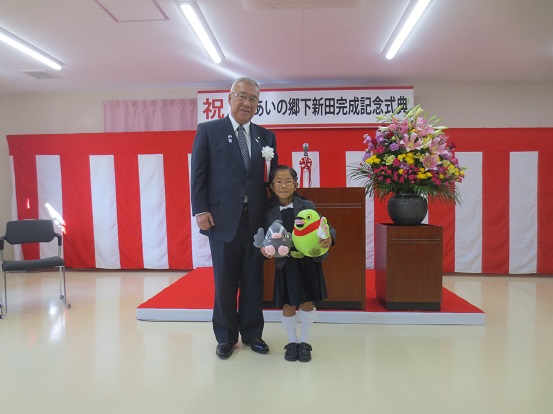 「祝 戸ヶ崎ふれあいひろば完成記念式典」と書かれた横幕と紅白幕が貼られた会場の演壇の前に、市長と紺色のワンピースを着てマスコットキャラクターかいちゃんとつぶちゃんのぬいぐるみを持っている新沼さんが並んでいる記念写真