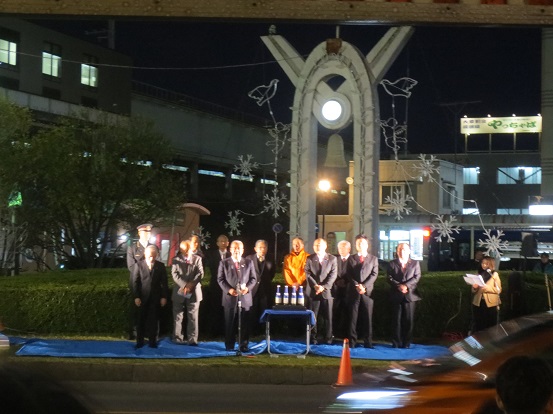 日が暮れて照明が照らされた駅前の小さな広場に敷かれたブルーシートの上に関係者が立ち、中央に立っている市長が挨拶を行っている様子の写真