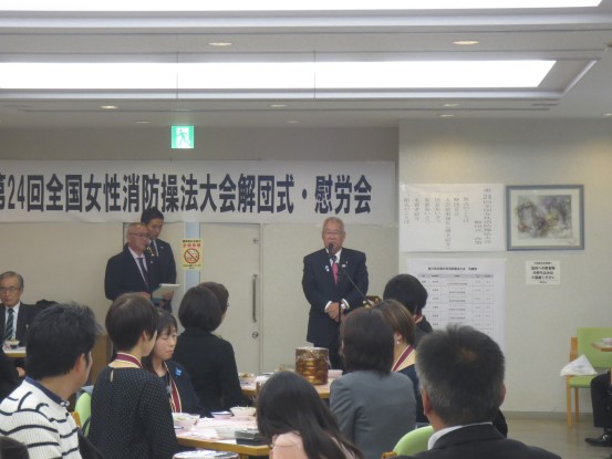 「第24回全国女性消防操法大会解団式・慰労会」と書かれた横幕が壁に貼られた室内で、手前の席に座っている参加者が奥に立つ市長の話を聞いている写真