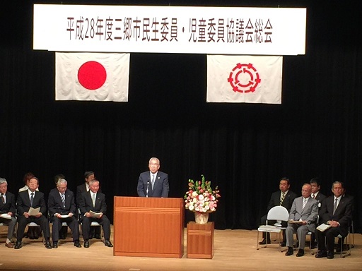 「平成28年度三郷市民生委員・児童委員協議会総会」と書かれた横断幕と国旗と三郷市旗が飾られたステージの両端に関係者が座り、中央の演壇で挨拶を行っている市長の写真