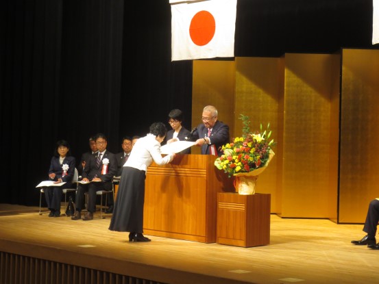 金屏風が立てかけられたステージの席に関係者が座り、中央の演壇を挟んで市長が白色のシャツを着た女性に感謝状の贈呈を行っている様子の写真