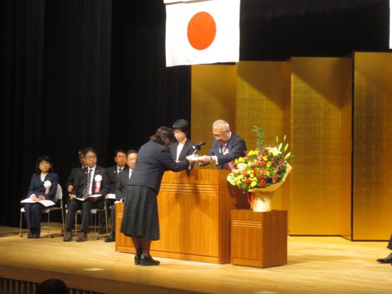金屏風が立てかけられたステージの席に関係者が座り、中央の演壇を挟んで市長が黒色のスカートスーツを着た女性に委嘱書の交付を行っている様子の写真