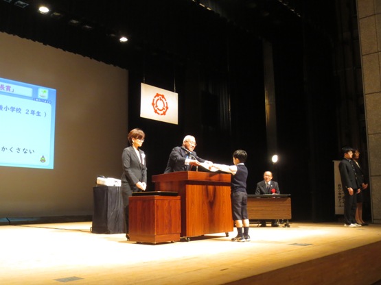 演壇を挟んで市長が向かい合っている男の子に賞状を手渡している表彰式の様子の写真