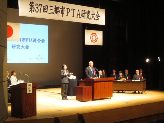 「第37回三郷市PTA研究大会」と書かれた横断幕やモニターが映されたステージの左右の席に関係者が座り、舞台中央で女性が手話を行う隣で挨拶を行っている市長の写真