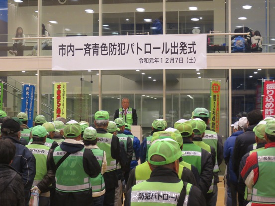 「市内一斉青色防犯パトロール出発式」と書かれた横断幕がガラス壁に貼られ、手前に設置された演壇に立ち挨拶を行っている市長の話を、向かい合って立っている緑色のキャップを被った方々が聞いている様子を真後ろから撮影した写真