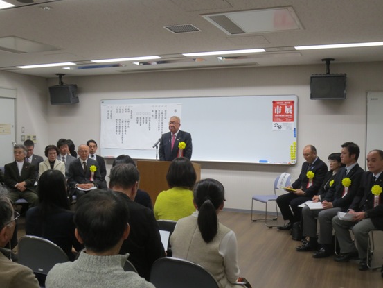 左右に設置された席に座っている関係者や手前の椅子に座っている参加者が、中央の演壇で挨拶を行っている市長の話を聞いている写真