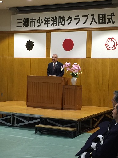 「三郷市少年消防クラブ入団式」と書かれた横幕と国旗が飾られた室内に設置された演壇で挨拶を行っている市長の写真