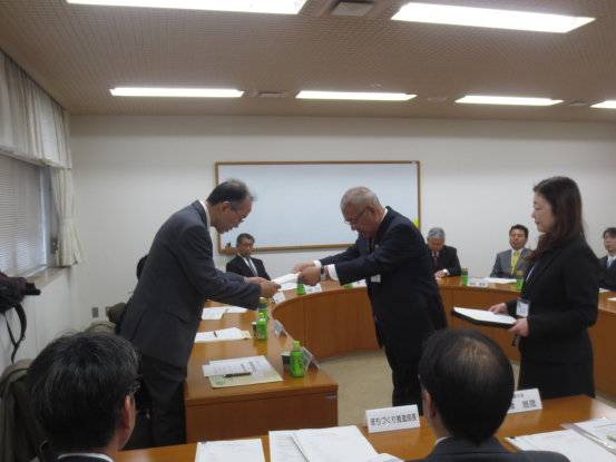 手前の席に関係者が座り、委員の方々がUの字の形の机の席に座り、左手前の男性が立ちあがって市長から委嘱書を受け取っている様子の写真