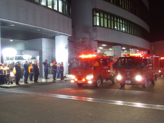 消防団員や市長が敬礼して見送るなか、ランプをつけた消防車が出陣していく様子の写真