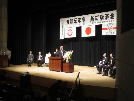 「令和元年度防災講演会」と書かれた横断幕が飾られたステージ左右の席に胸章リボンをつけた関係者が座り、中央の檀上で挨拶を行っている市長の写真