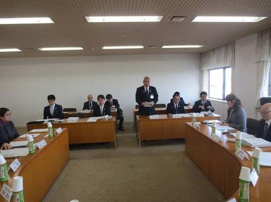 2列に設置された3名掛けの長テーブルの一番前に1人の男性が立ち、挨拶を行っている市長の話を聞いている関係者の方々を写した写真
