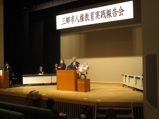 「三郷市人権教育実践報告会」と書かれた横断幕の舞台の左側の席に関係者が座り、ステージ左端に司会者が立ち、中央に手話通訳者の女性と挨拶を行っている市長を写した写真