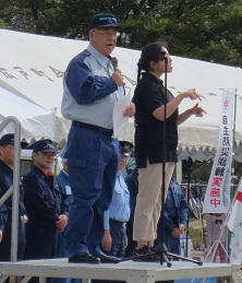 朝礼台の上で黒色のシャツを着た女性の手話と同時にマイクを持ち挨拶を行っている市長の写真