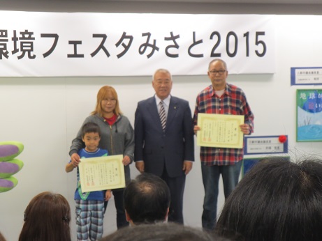 「環境フェスタみさと2015」と書かれた横幕の前で市長を挟んで男性と母親と男の子が賞状を持って並んで立っている写真