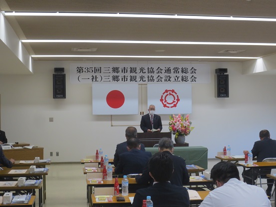 横断幕や国旗と三郷市旗が飾られた室内で、長机の席に座っている参加者が演壇で挨拶を行っている市長の話を聞いている後ろ姿の写真