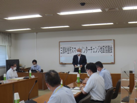 「三郷料金所スマートインターチェンジ地区協議会」と書かれた横幕が貼りつけられているホワイトボードの前の席に立ち挨拶の言葉を述べる三郷市長と、マイクとペットボトルのお茶が置かれている席に座り話を聞く関係者の写真
