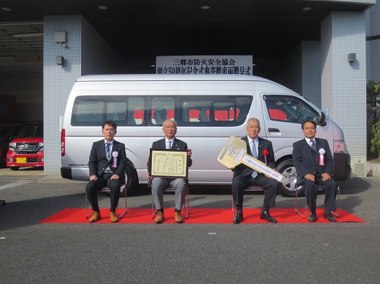 「三郷市防火安全協会創立50周年記念式典寄贈車両贈呈式」と書かれた横断幕と紅白幕が貼られ、トヨタのロゴが入った大きな鍵を持つ三郷市長と感謝状を持つ男性、関係者の男性2名がマイクロバスの前に敷かれたレッドカーペット上の椅子に座り記念撮影をしている写真
