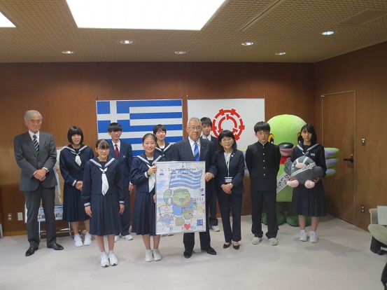 ギリシャ国旗と三郷市旗が吊るされている壁の前で、1名の女子中学生と三郷市長がギリシャ国旗を手に持ったかいちゃんと三郷市旗を手に持ったつぶちゃんが描かれている歓迎メッセージボードをともに持ち、関係者と中学生、かいちゃん・つぶちゃんとともに記念撮影をしている写真