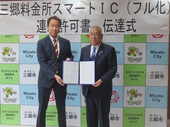 「三郷料金所スマートIC（フル化）連結許可書伝達式」と書かれたポスターが貼りつけられている三郷市のバックボードの前で、佐藤所長と三郷市長が連結許可書をともに持ち記念撮影をしている写真