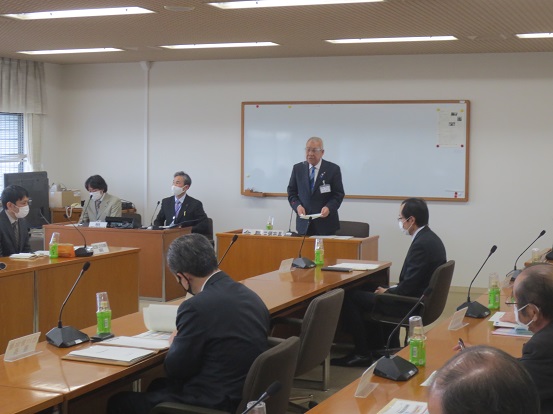 ホワイトボードの前の席に立ち挨拶の言葉を述べる三郷市長と、マイクと席札、お茶のペットボトルが置かれている席に座り話を聞く関係者の写真