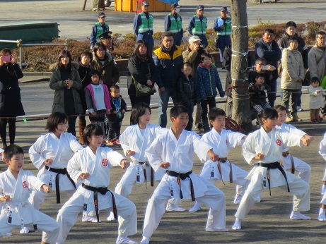 離れた後ろ側で観客が見ているなか、足袋を履き道着姿の子どもたちが両脚を大きく広げ正拳突きの構えで演武を行っている様子の写真
