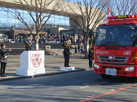 文化会館前の道路を走行している消防車に向かって、歩道に設置された檀上で敬礼を行っている市長の写真