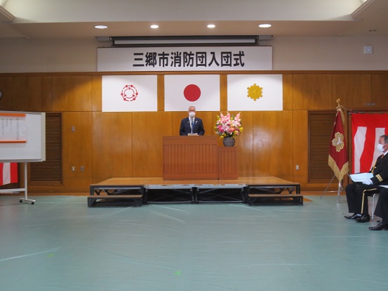 「三郷市消防団入団式」と書かれた横断幕と三郷市章、国旗、消防紋章が掲げられた会場で挨拶している三郷市長と、関係者の方々が右脇に座って聞いている写真