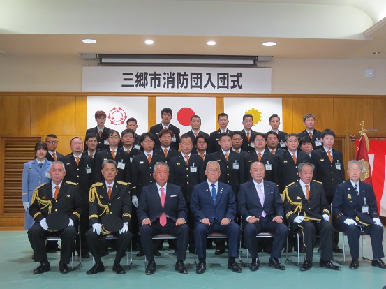 三郷市消防団入団式に参加された関係者の方々が横4列に並び最前列は椅子に座り、後列は立って一緒に記念撮影をしている写真