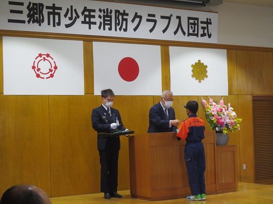 「三郷市少年消防クラブ入団式」と書かれた横断幕と三郷市章、国旗、消防紋章が掲げられた会場で、三郷市長がオレンジとネイビーの制服を着た男の子に少年消防手帳及びクラブ印章を交付している様子の写真
