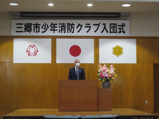 「三郷市少年消防クラブ入団式」と書かれた横断幕と三郷市章、国旗、消防紋章が掲げられた会場で、挨拶している三郷市長の写真