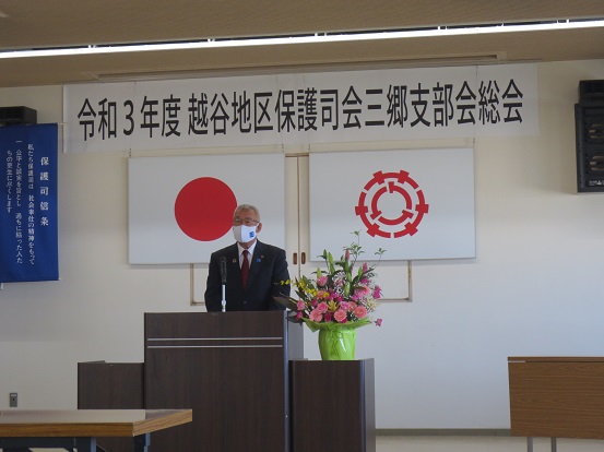 「令和3年度越谷地区保護司会三郷支部会総会」と書かれた横断幕と国旗、三郷市章が飾られた会場で、ピンクや黄色の花が飾られた演台に立って挨拶する三郷市長の写真