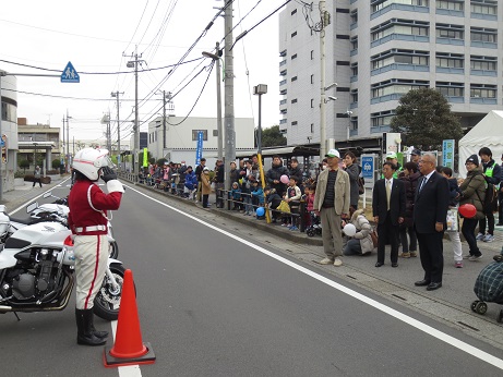 道路に停めた白バイの傍でヘルメットを被り赤と白の制服を着用した白バイ隊員が歩道に立っている市長に向かって敬礼を行っている写真