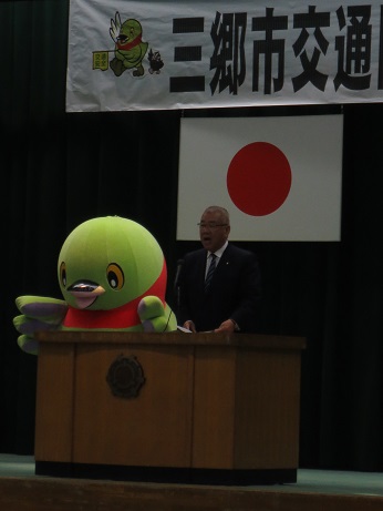 「三郷市交通防犯フェア」と書かれた横断幕の下、マスコットキャラクターかいちゃんの右側の演壇で挨拶を行っている市長の写真