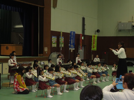 ステージ下で鍵盤ハーモニカや木琴などの楽器を先生の指揮に合わせて演奏を行っている園児の写真